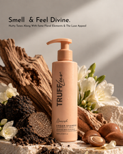 TRUFFluv Nourish Argan Shampoo bottle in a warm beige tone, styled with natural wood driftwood, white flowers, and argan nuts on a stone surface. Text at the top reads 'Smell & Feel Divine.