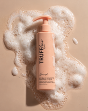 A bottle of Trufflerush Nushush Argan Shampoo with a peach-colored pump, placed on a beige surface surrounded by white foam.