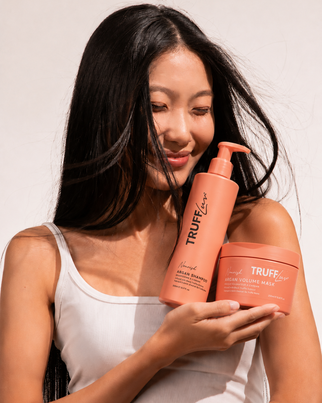 A woman with long black hair holds two Truff Love haircare products, including a peach-colored shampoo bottle and a matching volume wrap jar, against a clean white background.