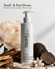 A bottle of TruffLeau Indulge Truffle Shampoo stands on a stone surface with white flowers, black truffles, and wood around it. Text at the top reads, 'Smell & Feel Divine.